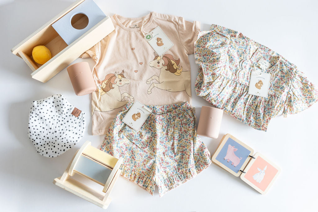 Flat lay of a SnuggleBox Infant (12-24 Months) collection featuring a unicorn-print top, floral shorts, a ruffled floral top, wooden play toys, and a polka-dot bib.