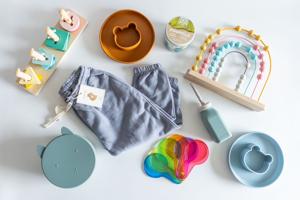 Flat lay of a SnuggleBox Toddler (24-36 Months) collection, featuring soft grey sweatpants, bear-shaped silicone plates, a blue water bottle, a wooden rainbow abacus, and colorful measuring spoons.