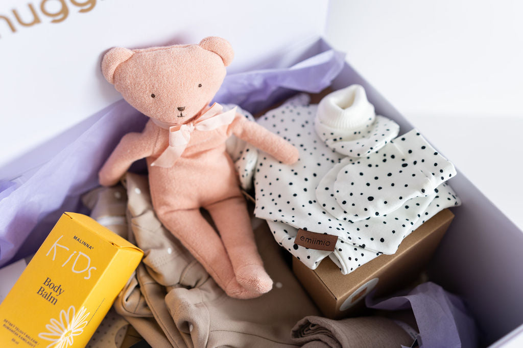 Baby items including a pink teddy bear, baby clothes, and a yellow box in a box with purple tissue paper.