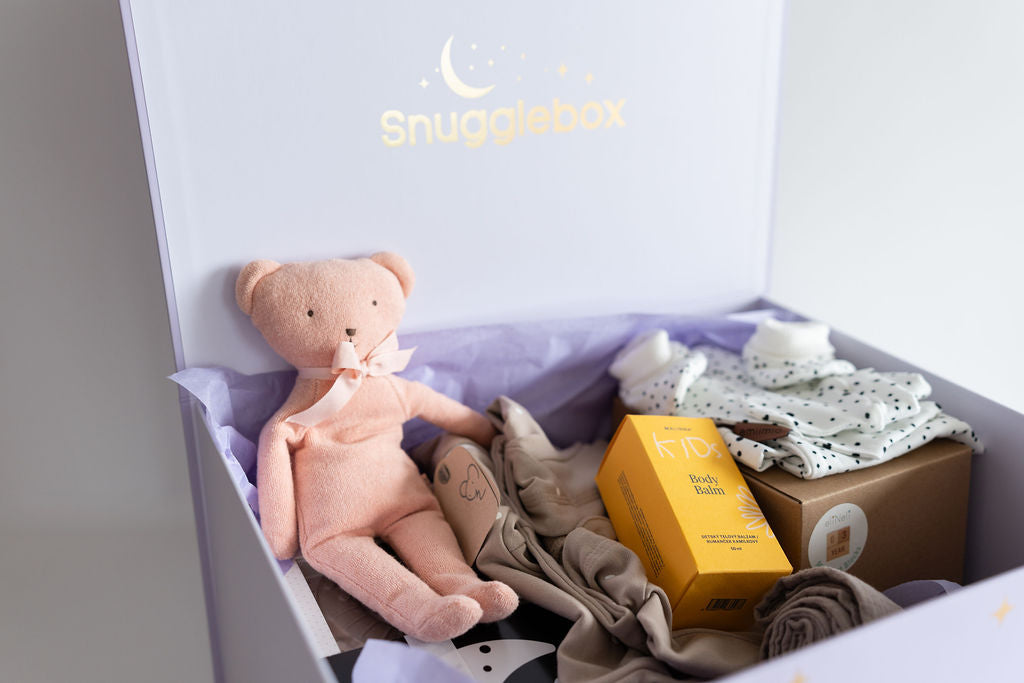 Open SnuggleBox revealing a pink plush bear, baby body balm, and folded baby clothing, showcasing the curated surprise and quality.
