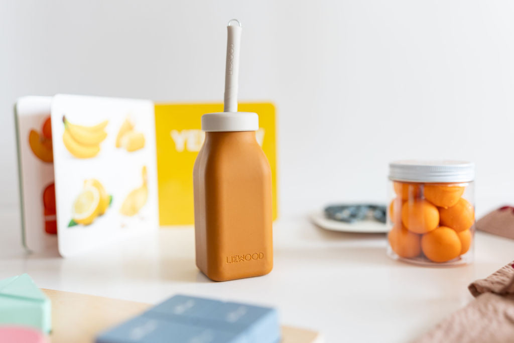 Brown bottle with a straw on a white surface with books and a jar of oranges in the background
